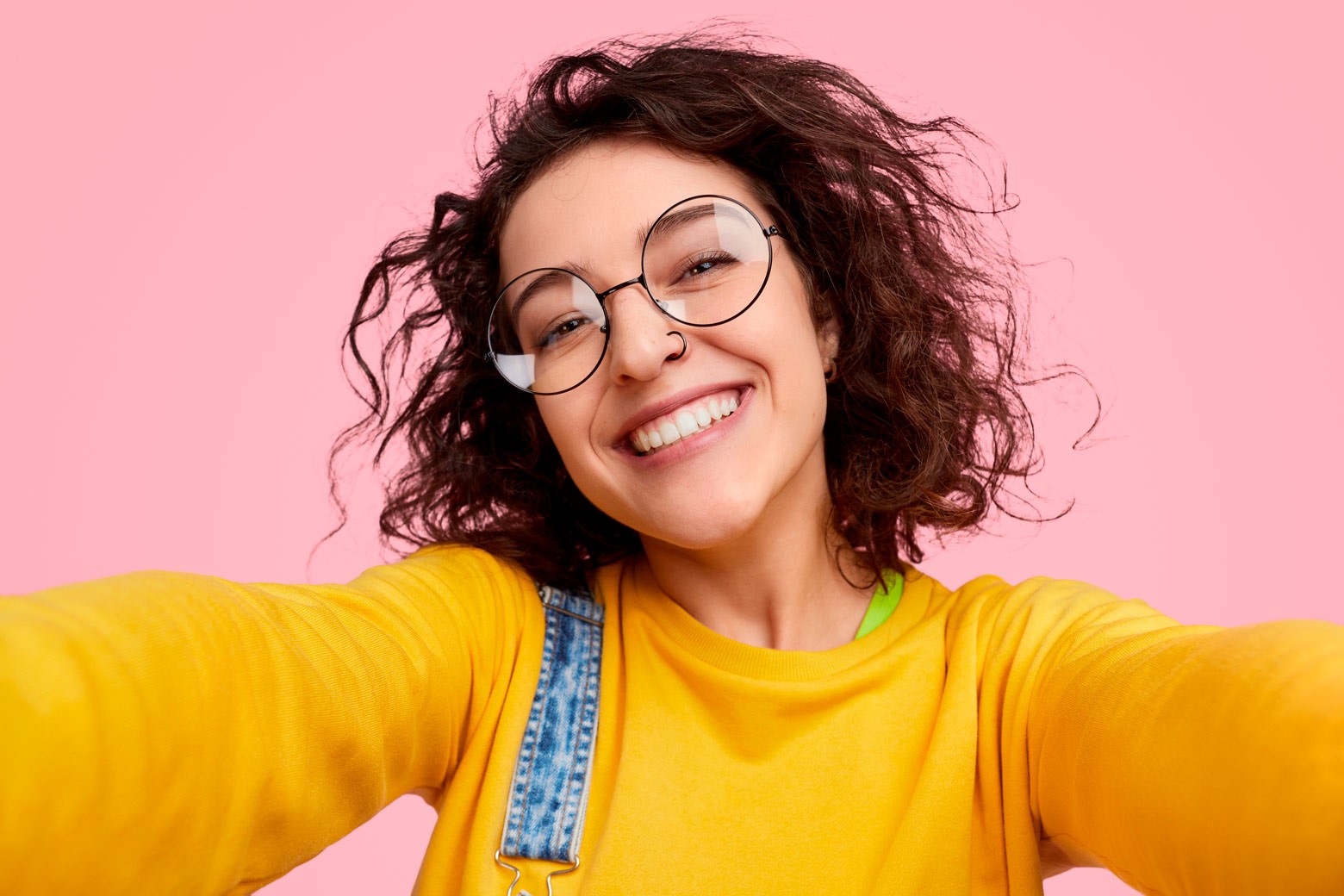 A woman with glasses is capturing her new look in a selfie on a pink background.