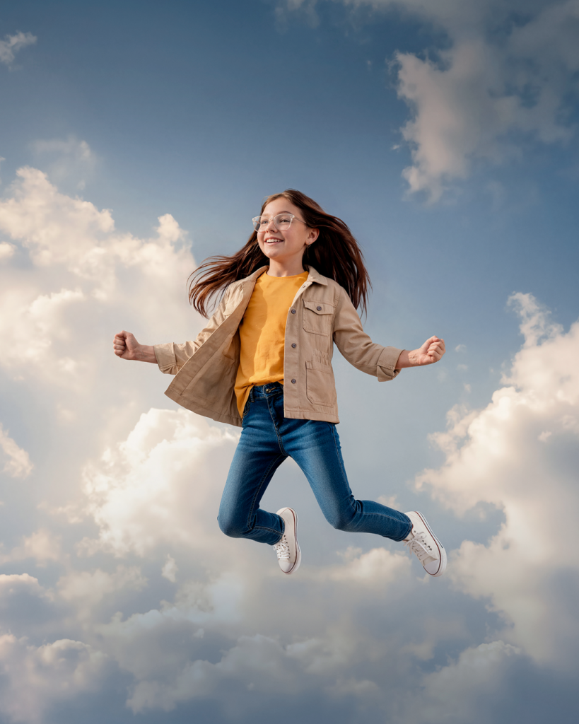 A girl wearing glasses, a tan jacket, yellow shirt, jeans, and white sneakers jumps in the air against a cloudy sky.