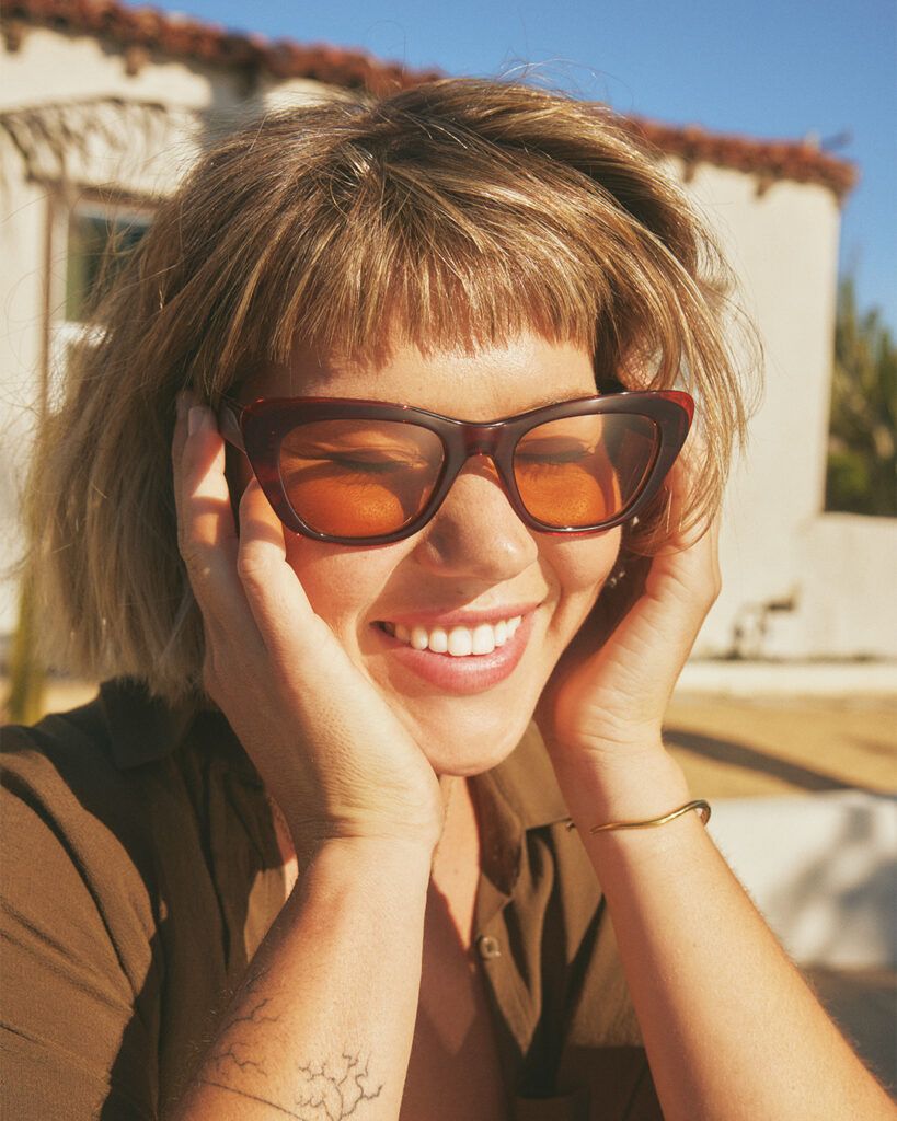 Woman smiling with sunglasses, holding her face with both hands.