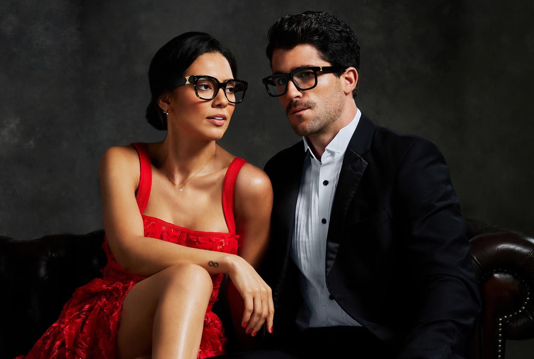 A woman in a red dress and a man in a black tuxedo sit side by side on a dark couch, both wearing black glasses and looking slightly away from the camera.