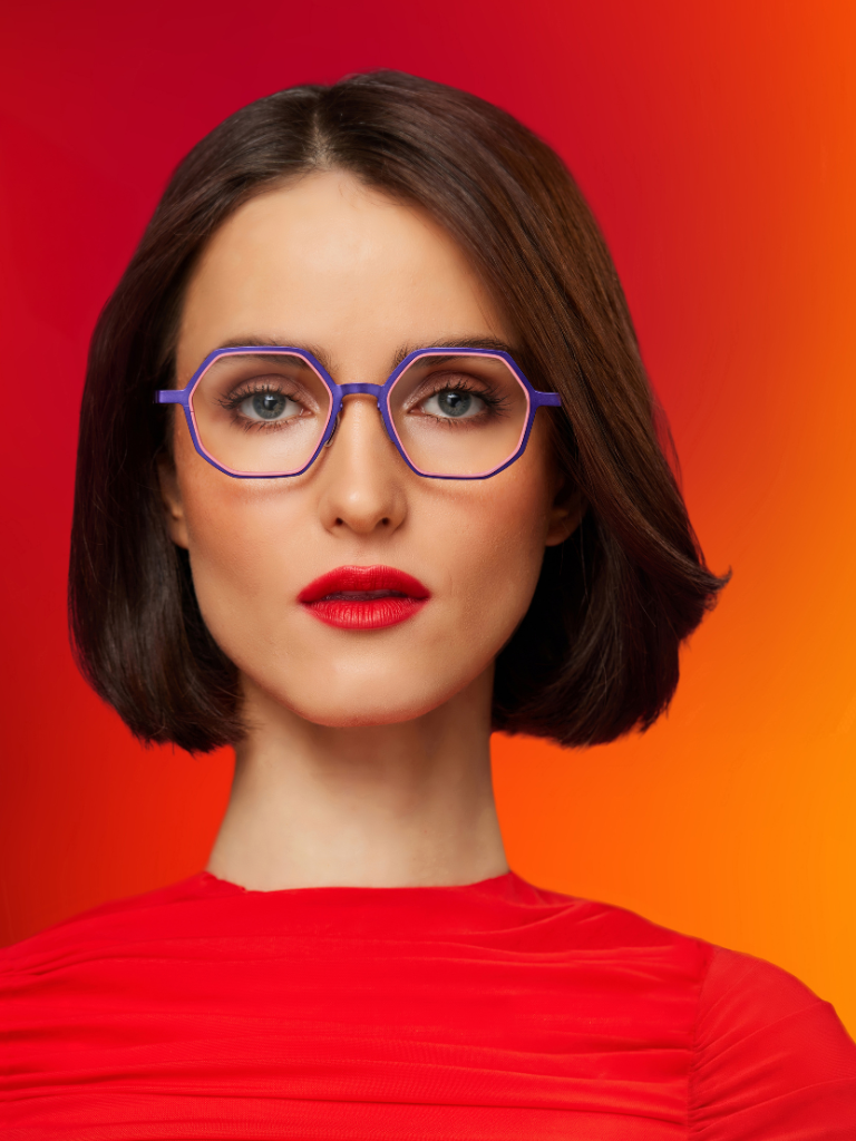Woman with short dark hair, wearing purple glasses and a red top, posing against a vibrant red and orange gradient background.