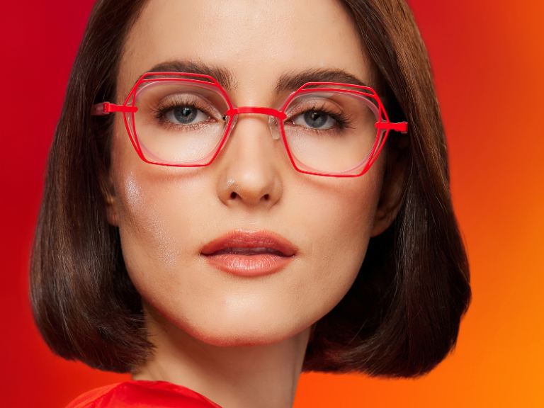 Woman with straight brown hair wearing red glasses and top, against a red-orange background. Text reads, 