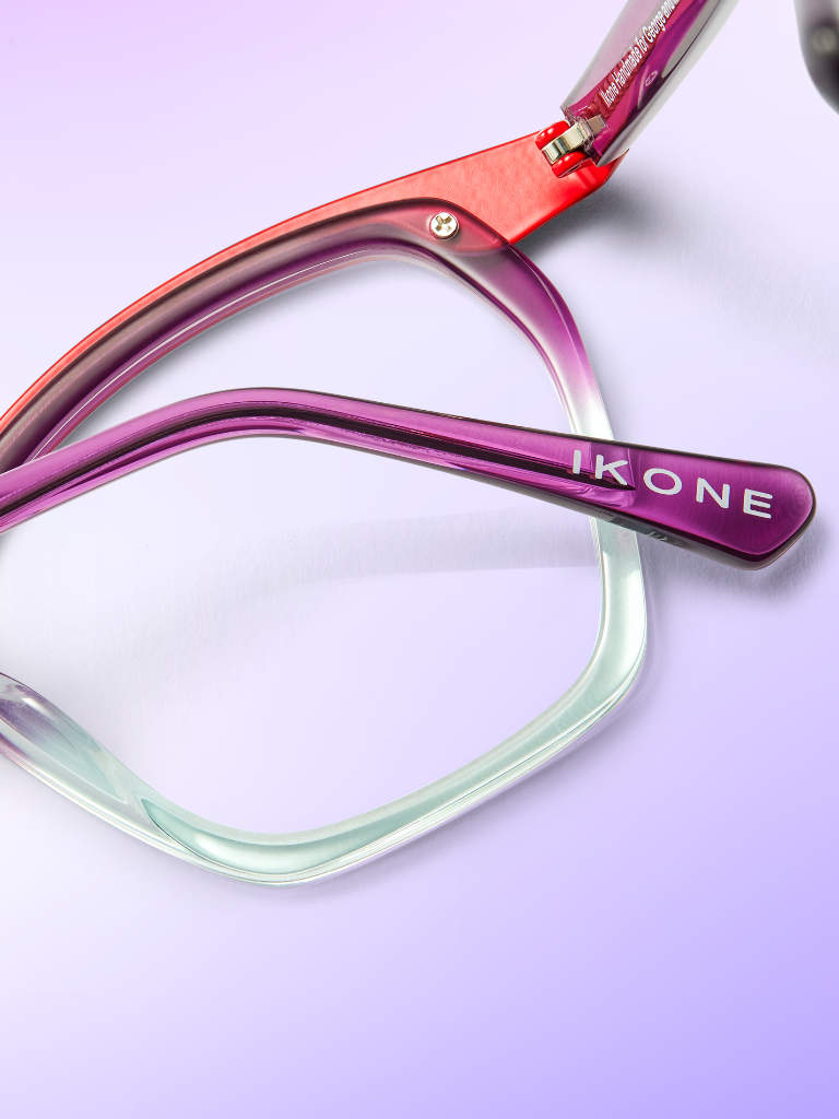 Close-up of a pair of purple and mint green eyeglass frames labeled “IKONE” on the temple, set against a soft purple gradient background.